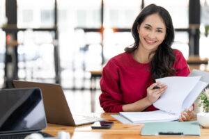 Geschäftsfrau arbeitet mit Papierkram und Laptop-Computer, um Geschäftsdaten und Finanzen im Büro zu analysieren, lächelnd und in die Kamera schauend.