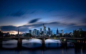 Die Skyline von Frankfurt bei Nacht, mit beleuchteten Wolkenkratzern und einer Brücke über den Main, die den nächtlichen Himmel reflektiert.
