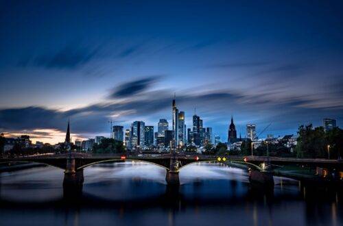 Die Skyline von Frankfurt bei Nacht, mit beleuchteten Wolkenkratzern und einer Brücke über den Main, die den nächtlichen Himmel reflektiert.