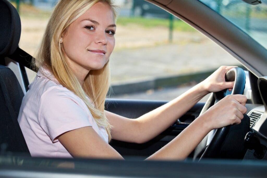 Junge blonde Frau sitzt lächelnd am Steuer eines Autos und blickt in die Kamera