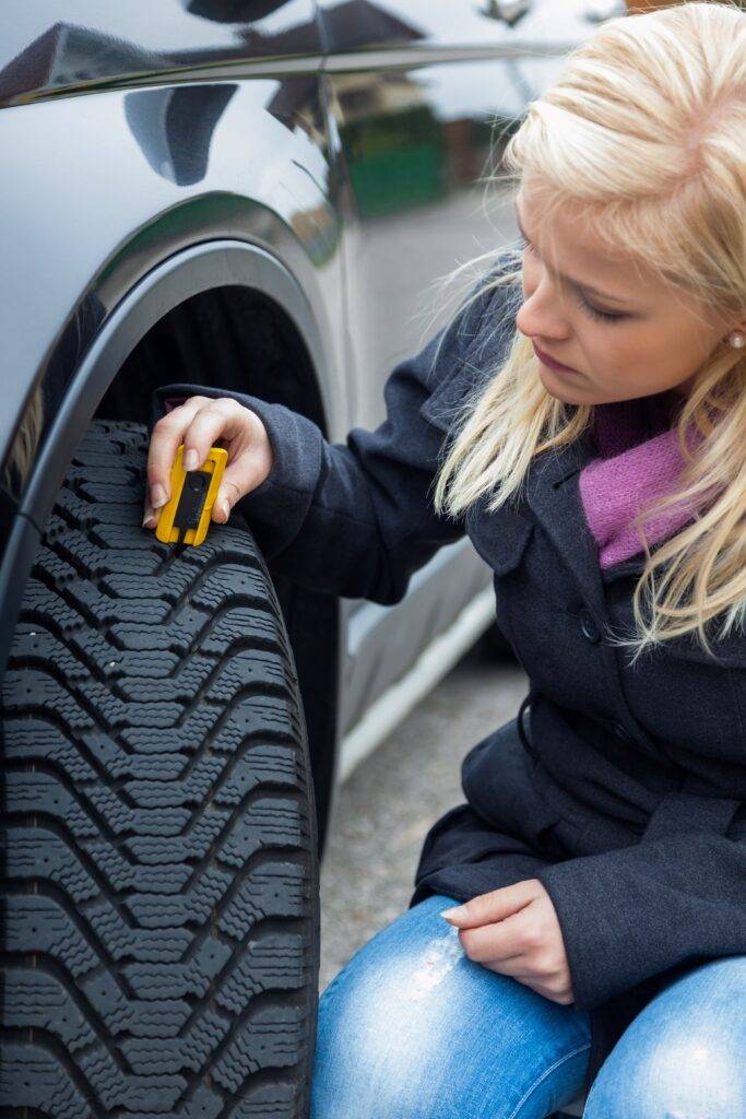 Junge Frau in einer schwarzen Jacke misst die Profiltiefe eines Autoreifens mit einem Messgerät.