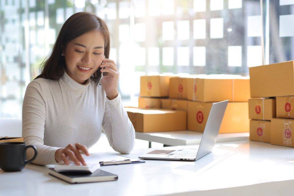 Eine Geschäftsfrau spricht am Telefon und arbeitet an ihrem Laptop in einem Büro mit Paketen im Hintergrund, unterstützt durch eine Amazon Agentur, um ihre E-Commerce-Aktivitäten zu optimieren.