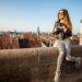 Frau in gestreifter Hose und Lederjacke hält eine Kamera und sitzt auf einer Mauer mit Blick auf die Altstadt von Nürnberg bei sonnigem Wetter.