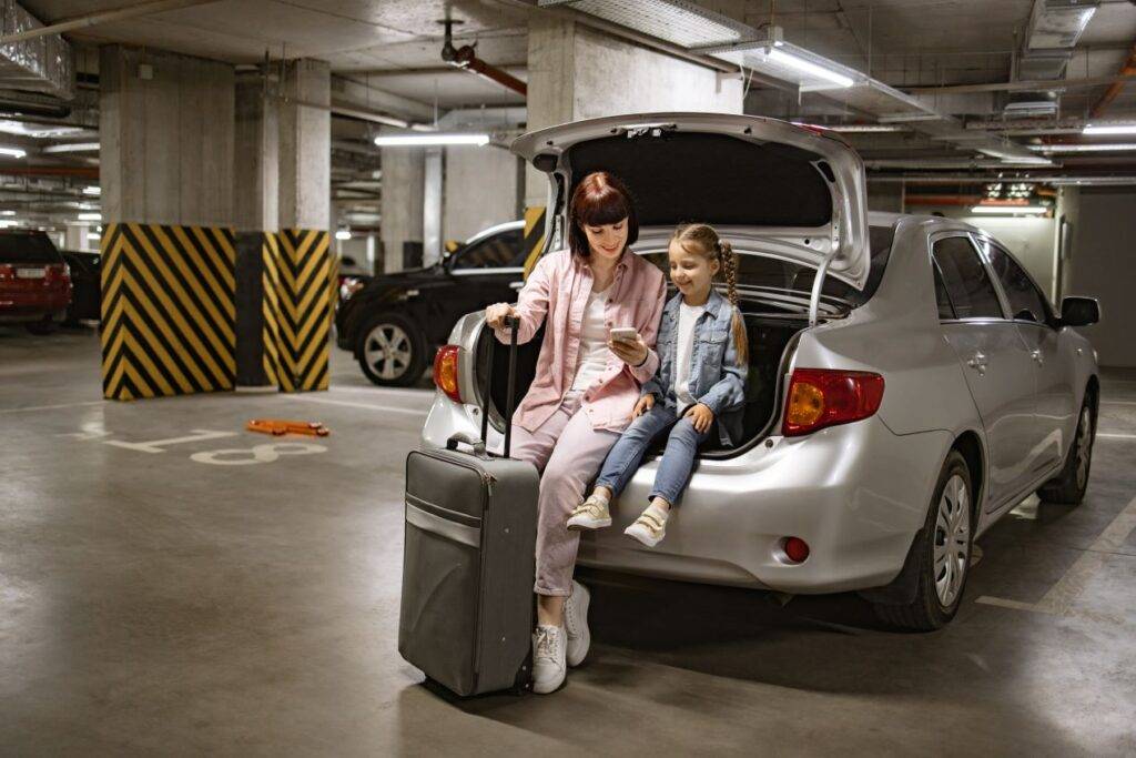 Mutter und Tochter sitzen im Kofferraum ihres Autos in einem Parkhaus, bereit zum Abflug, mit einem Koffer und Handy. Perfektes Beispiel für günstig parken am Flughafen Nürnberg.