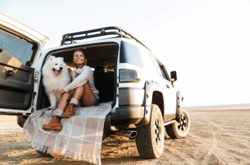 Junge Frau sitzt mit ihrem weißen Hund im geöffneten Kofferraum eines Geländewagens am Strand und genießt die Freiheit.