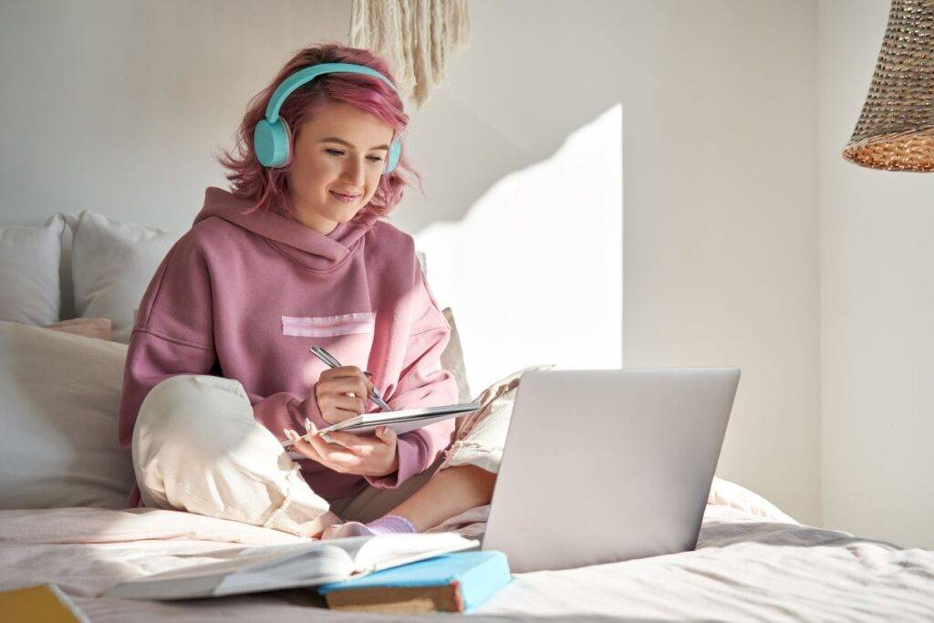 Junge Frau mit rosa Haaren und blauen Kopfhörern sitzt auf dem Bett und arbeitet konzentriert an ihrem Laptop, während sie Notizen macht.