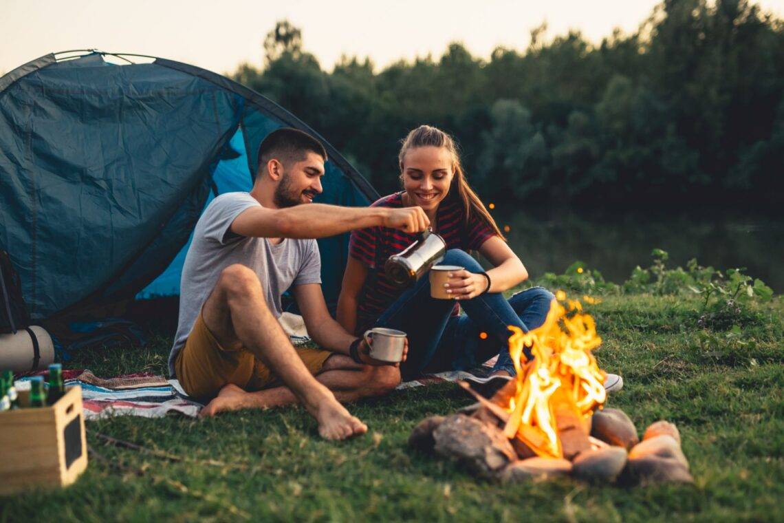 Zwei Personen sitzen bei Sonnenuntergang vor einem Zelt am Lagerfeuer und genießen Heißgetränke, ideal für entspannte Outdoor-Abenteuer.