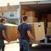 Zwei Maenner tragen Umzugskartons in einen Transporter vor einem Wohnhaus in der Sonne