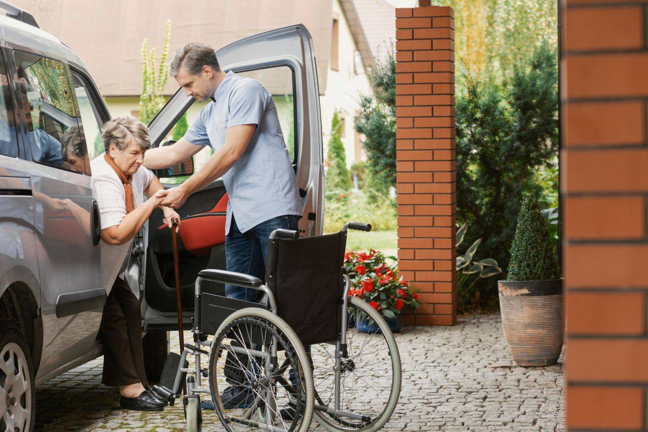 Junger Mann hilft Seniorin vom Auto in den Rollstuhl – praktisches Arbeitsfeld fuer Alltagsbegleiter Quereinsteiger im mobilen Alltag