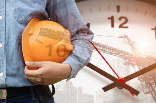 Bauarbeiter mit Schutzhelm vor Uhr und Kran bei Zeitdruck