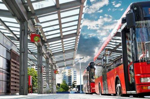 Rote Linienbusse am modernen Busbahnhof unter Glasdach mit Stadtgebäuden im Hintergrund, Symbol für ÖPNV Jobs.